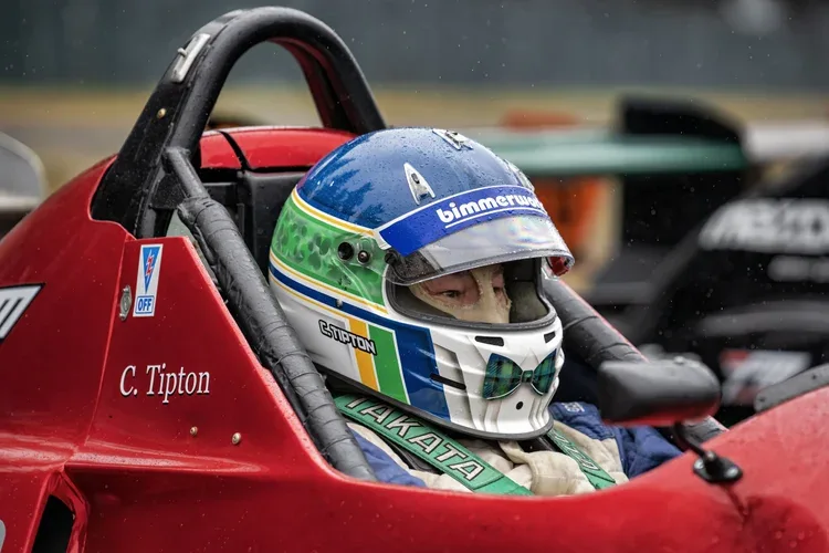 Close-up of a race car driver sitting in a red racing cockpit, wearing a blue, green, yellow, and white helmet with the text 'bimmerworld' on it, rain droplets on the helmet, and a focused expression visible through the helmet visor.