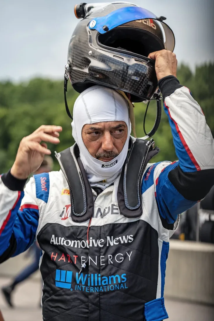 A race car driver is preparing for a race, putting on a black and blue racing helmet. The driver is dressed in a racing suit with sponsor logos including 'Automotive News,' 'Harte Energy,' and 'Williams International.' The background is blurred with