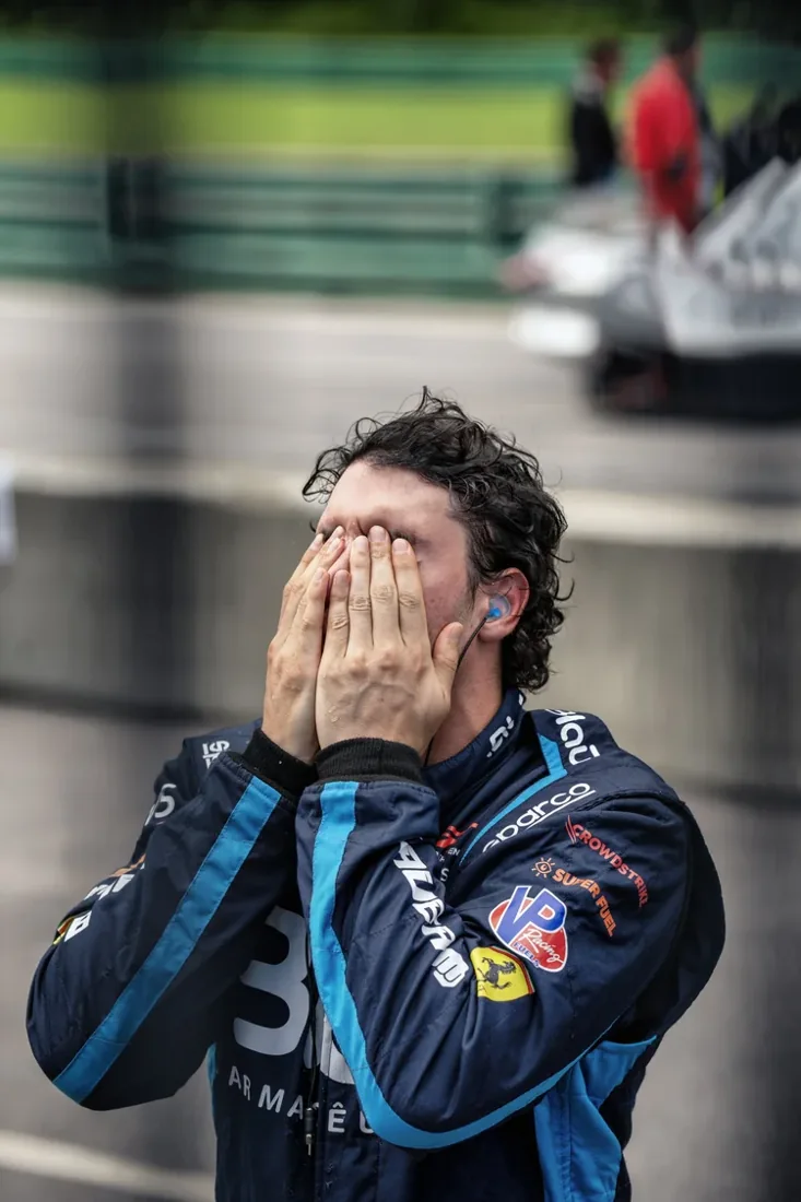 A race car driver with dark curly hair, wearing a racing suit with various sponsor logos, covering his face with his hands, in front of a blurred background showing part of a race track and some people in the background.