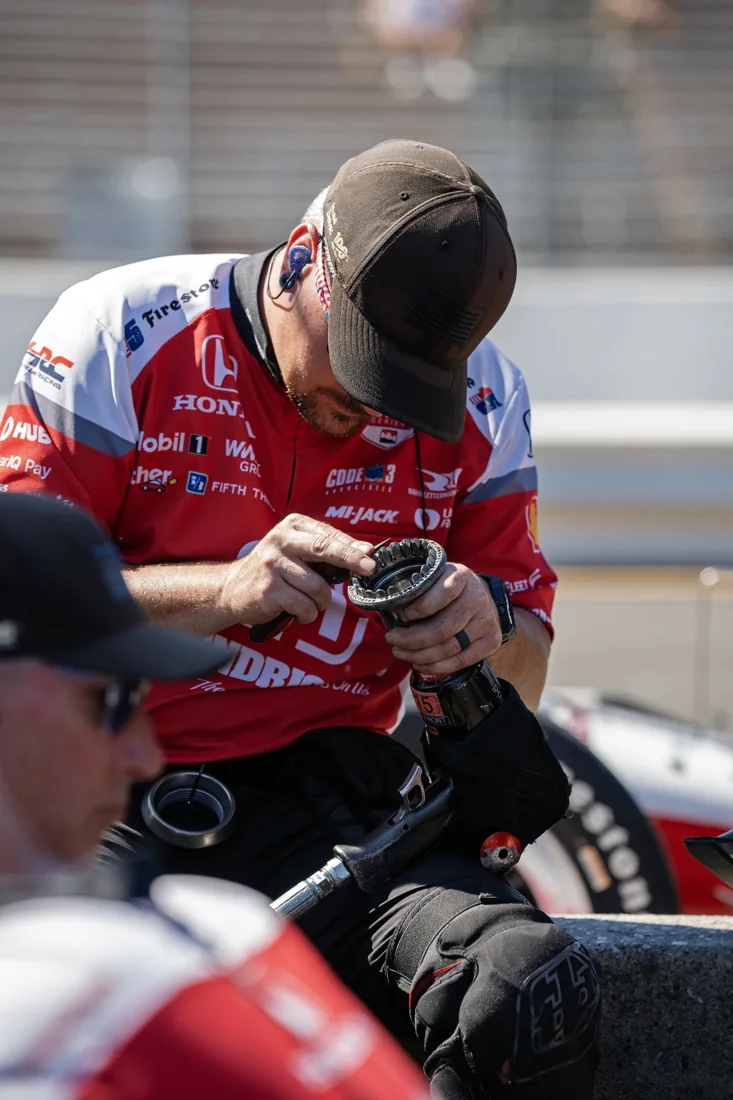 A male race car driver in a red and white racing suit and black cap is handling a motorcycle part or tool, with other individuals and motorcycles in the background.
