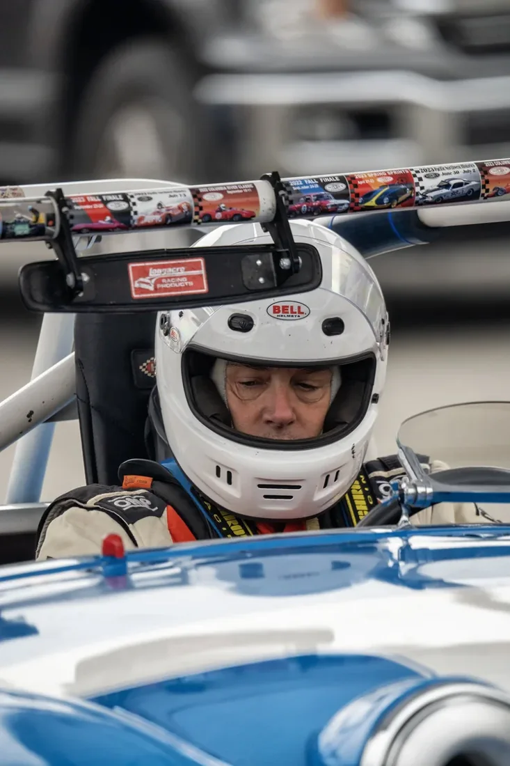 Close-up of a race car driver wearing a white helmet with a Bell logo, seated inside a racing vehicle. The background shows a blurred image of race cars and a metal structure with an information strip displaying images and details of race cars and ra