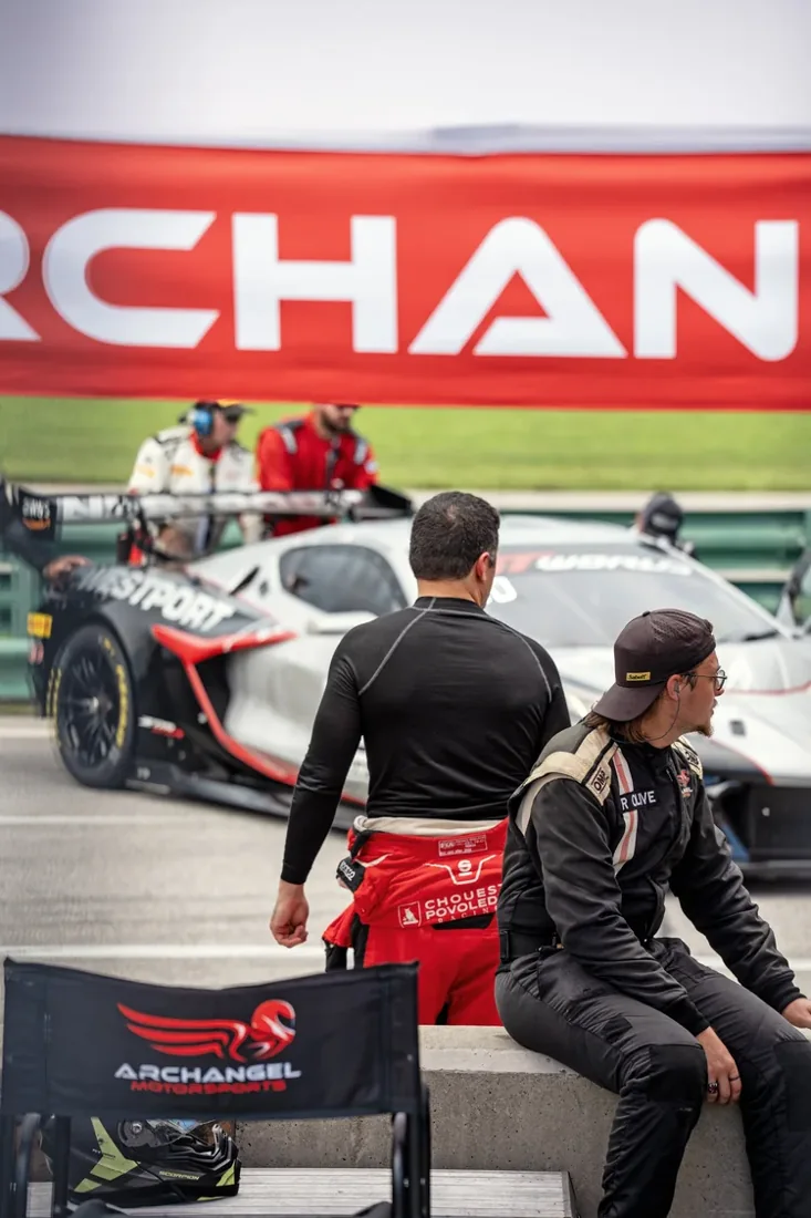 Race car drivers sitting and standing near race cars in a pit lane, with team members and crew around. A banner with the word 'ORCHAN' is visible in the background.