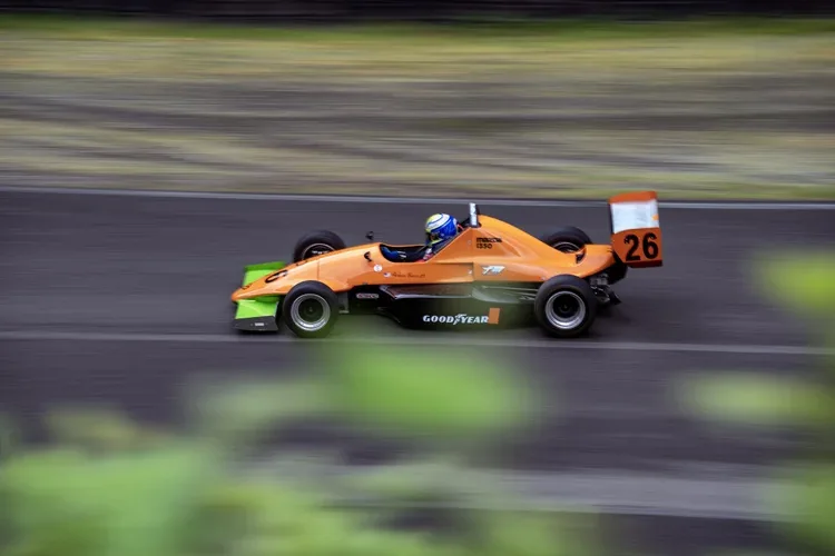 A bright orange race car with black and green accents speeding on a race track. The driver is wearing a blue helmet with yellow and white details. The car has the number 26 on the rear wing and various sponsorship logos, including Goodyear, on the si