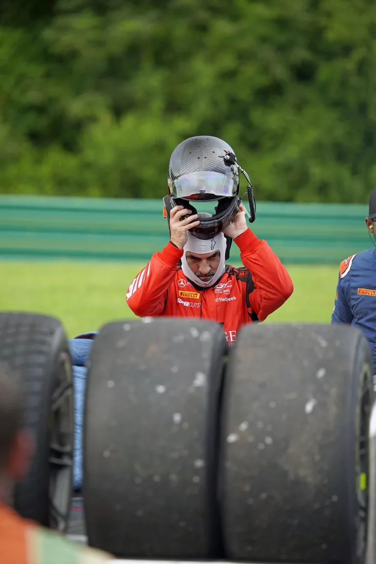 Race car driver in red racing suit holding helmet, seen from behind behind race car tires.