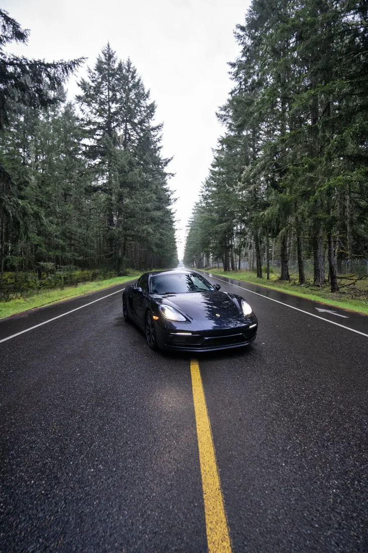 A black sports car on an empty, wet road surrounded by tall pine trees on both sides, on a cloudy day.