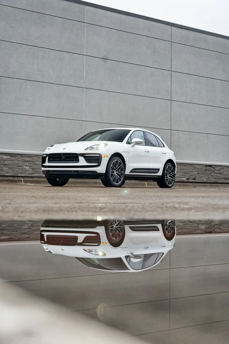 A white Porsche SUV parked in front of a large gray building with horizontal panels. The photo shows both the car and its mirror reflection on the surface below.