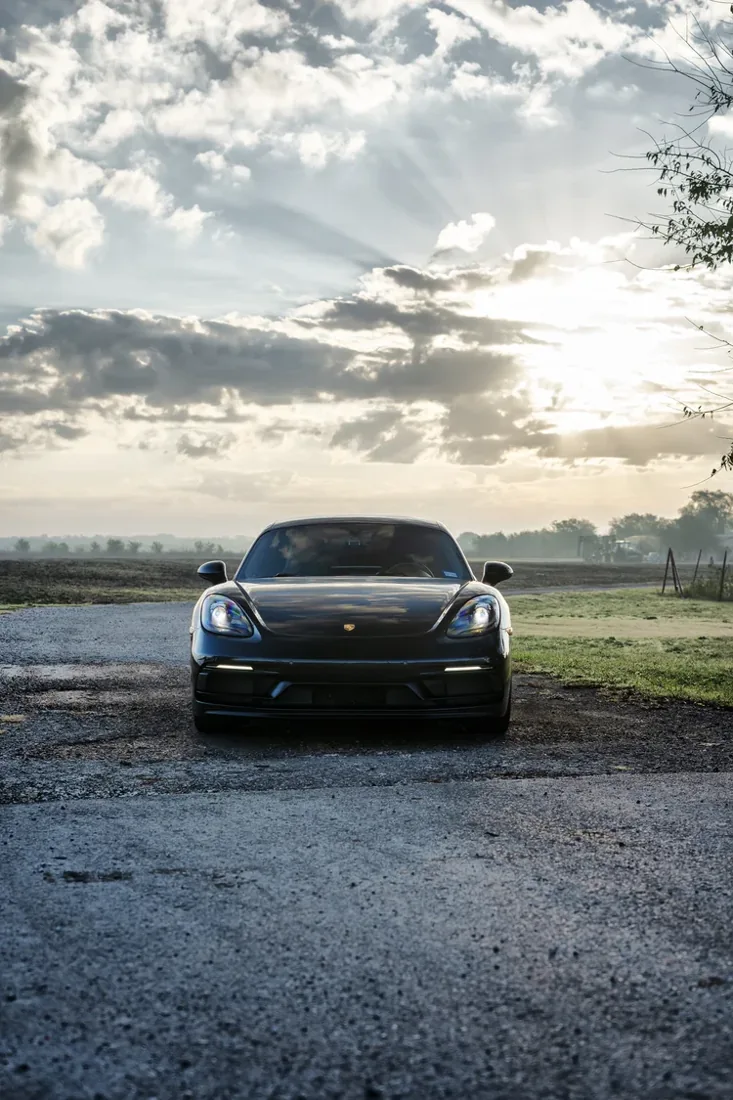 A black sports car parked on a gravel surface with a cloudy sky and sunshine behind it.