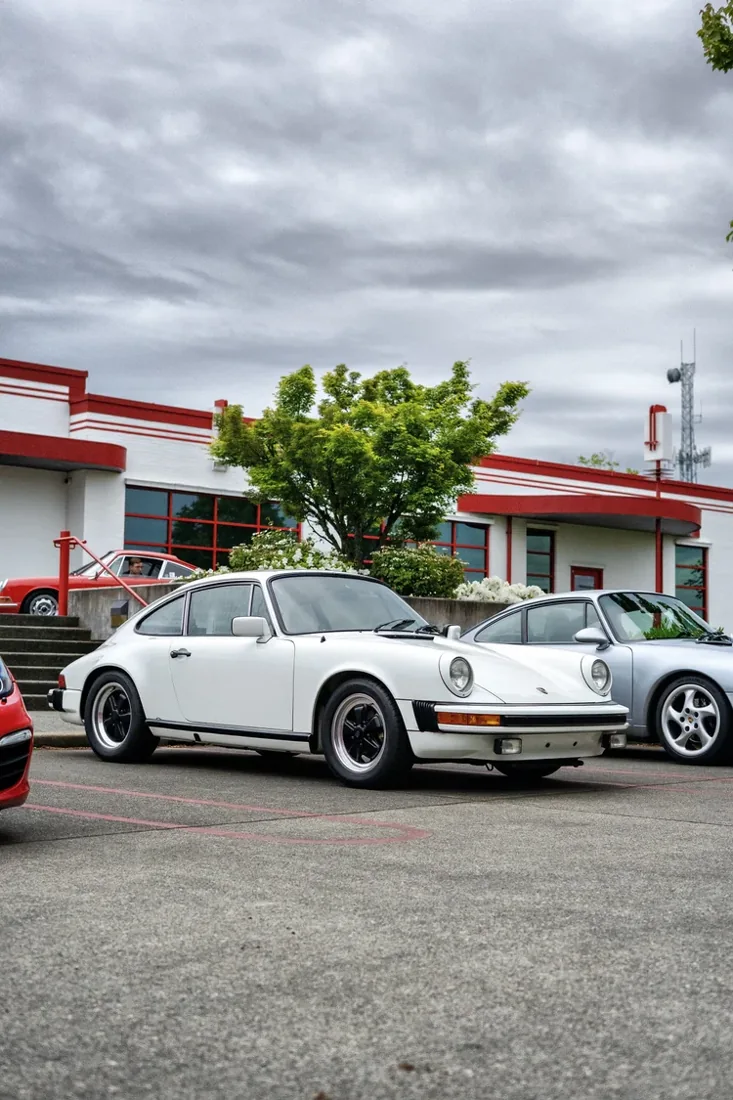 A white classic Porsche 911 parked in a parking lot in front of a building with red accents and a small green tree under a cloudy sky.