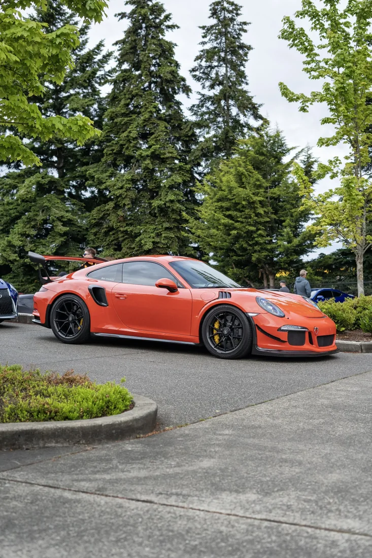 A red sports car parked in a parking lot surrounded by trees and other vehicles, with a few people standing nearby.