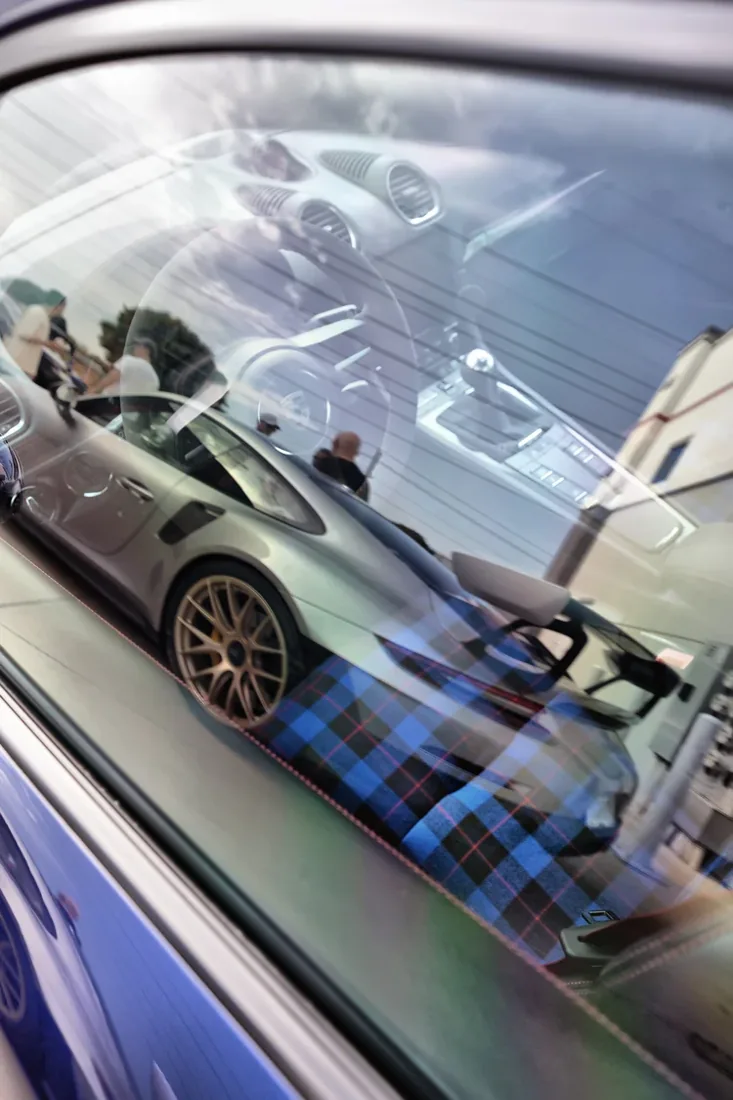 Reflection of a silver sports car with a checked blue and red interior from inside a car with a partially visible window. The photo shows the dashboard, steering wheel, and passenger seat through the window, with a cloudy sky and some people in the b