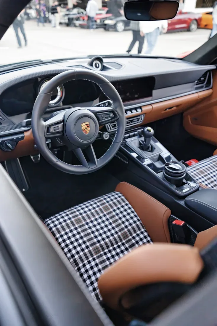 The interior of a Porsche vehicle showing the steering wheel with the Porsche logo, dashboard, gear shifter, cup holder, and front seat with a checkered and brown upholstery.