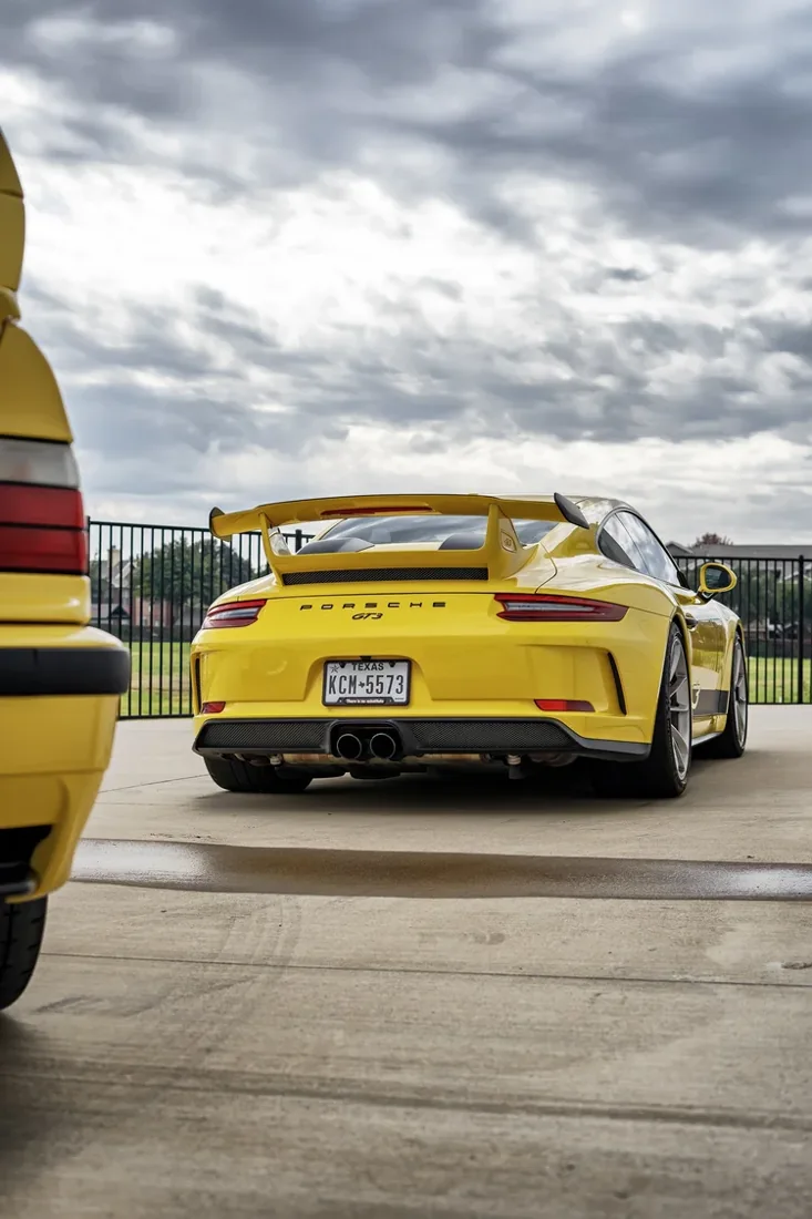 A yellow Porsche sports car with a large rear spoiler parked behind a black fence, with another yellow vehicle partially visible on the left side of the image. The background shows a cloudy sky and some trees.