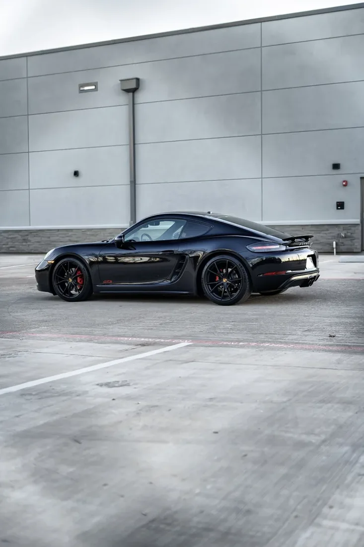 A black Porsche sports car parked in an empty parking lot against a gray concrete building with minimal design.