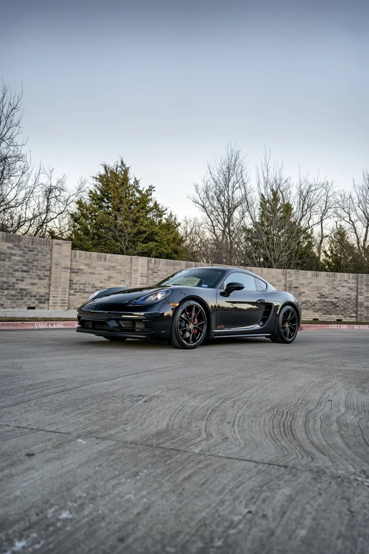 A black sports car parked on a concrete parking lot with a brick wall behind it. The background features leafless trees and a clear sky.
