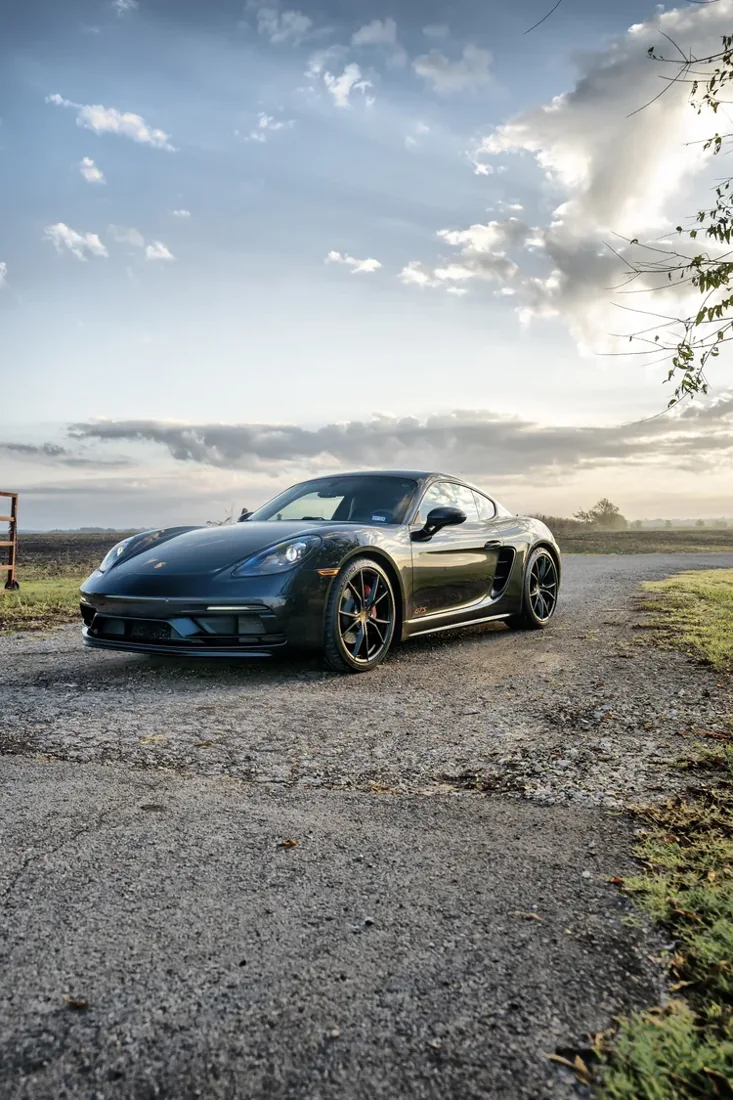 Black sports car parked on a gravel surface with a rural landscape and cloudy sky in the background.