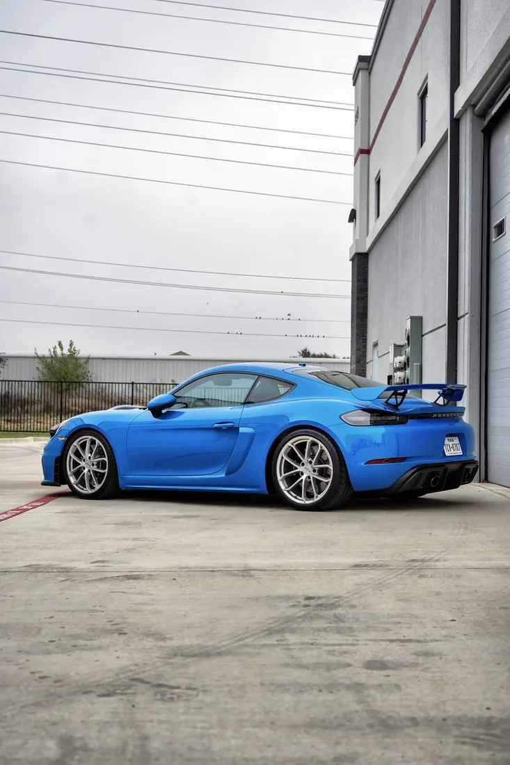 A bright blue Porsche sports car parked on a concrete lot next to a beige building with multiple windows and a garage door.
