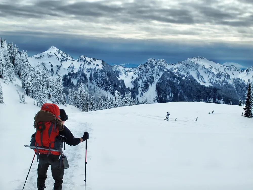 A person dressed in winter gear, carrying a backpack and using trekking poles, hiking on a snowy trail through a mountainous landscape with snow-covered trees and rugged peaks under a cloudy sky.