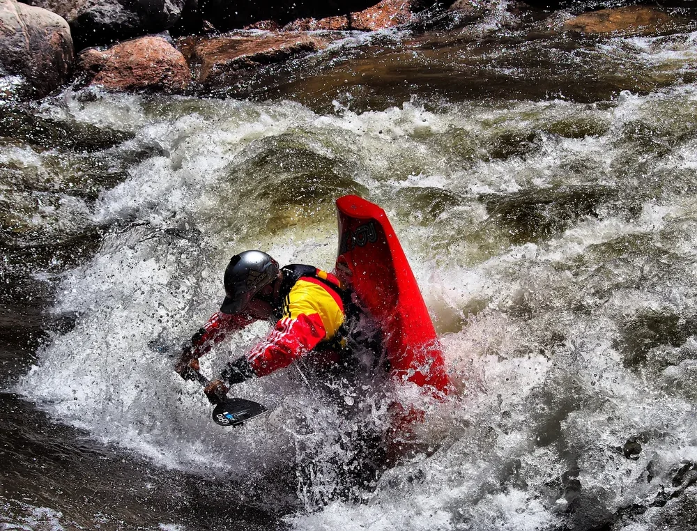 A person in a red kayak wearing a helmet is navigating through white water rapids.