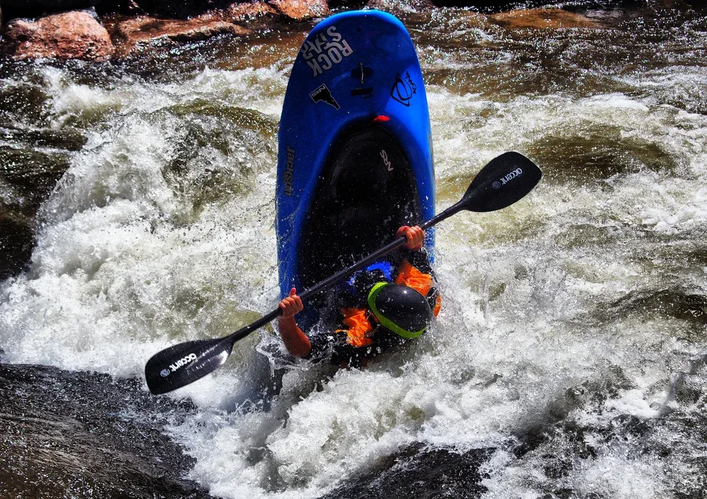 Person kayaking in rough water with foam, wearing a helmet and orange lifejacket, holding a paddle.
